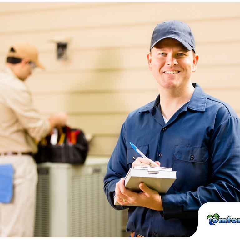 A smiling technician in a blue uniform holding a clipboard, with another technician working in the background.