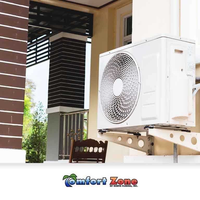 An air conditioning unit installed on a bracket outside a modern house, with the "comfort zone" logo visible at the bottom left.