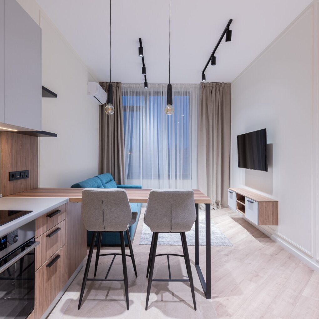 Modern kitchen with a dining area featuring a wooden table, two grey chairs, pendant lights, and integrated appliances, leading to a cozy living space with a tv.